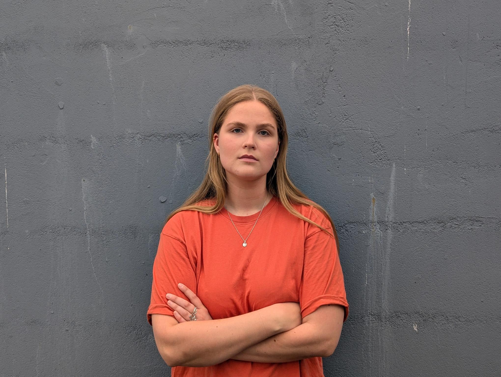 A young woman in an orange t-shirt with crossed arms stands against a grey wall.