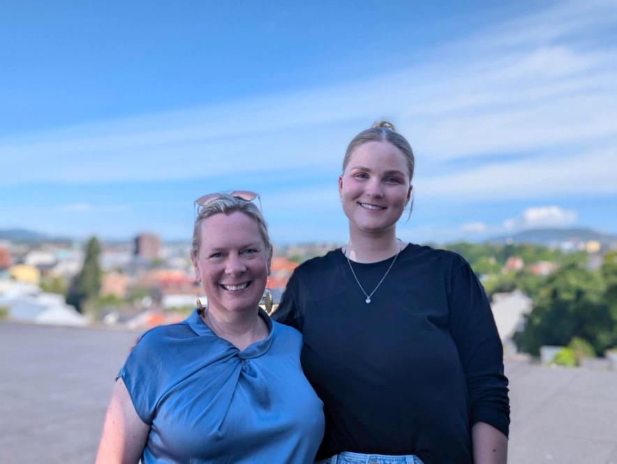 Two smiling women stand outdoors with a city skyline and blue sky in the background.