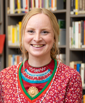 a woman in a red and green sweater smiles in front of a bookshelf