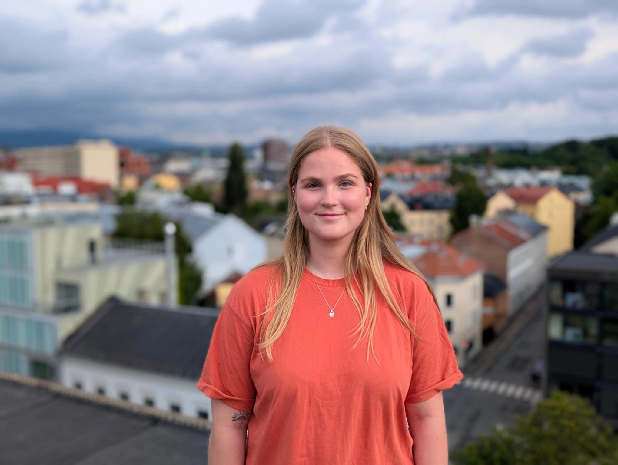 A smiling young woman with blonde hair and an orange shirt stands against a blurred city background.