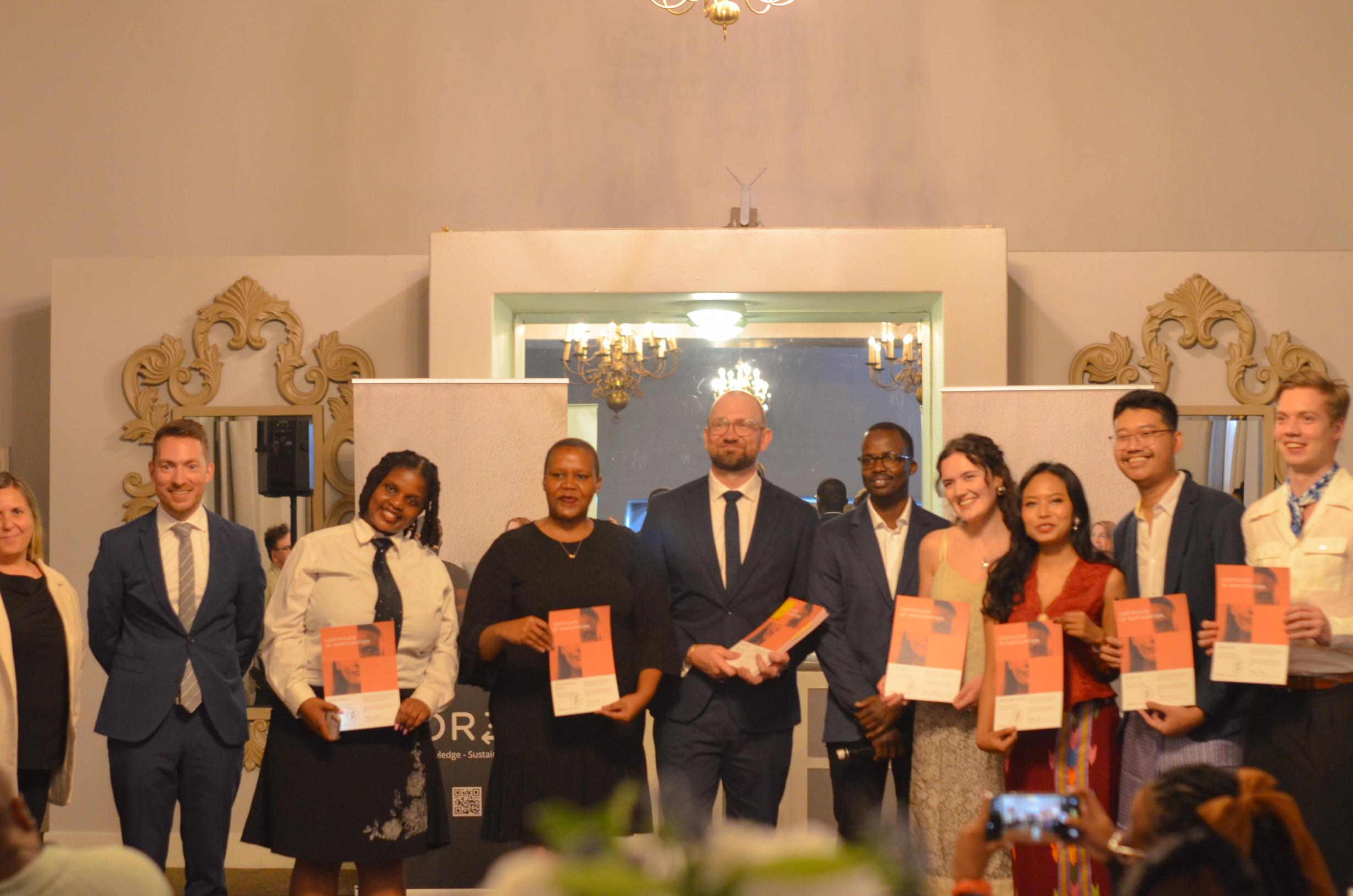 A diverse group of men and women in business attire stands together, holding certificates or awards at an indoor event.