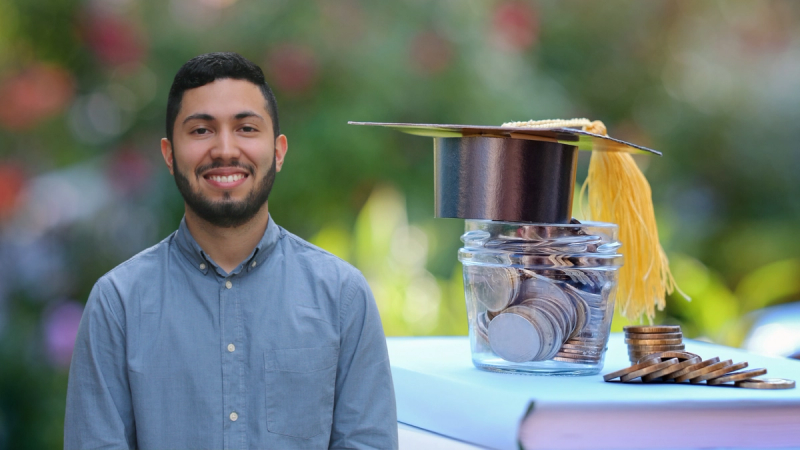 A man stands next to a jar of coins and a graduation cap.