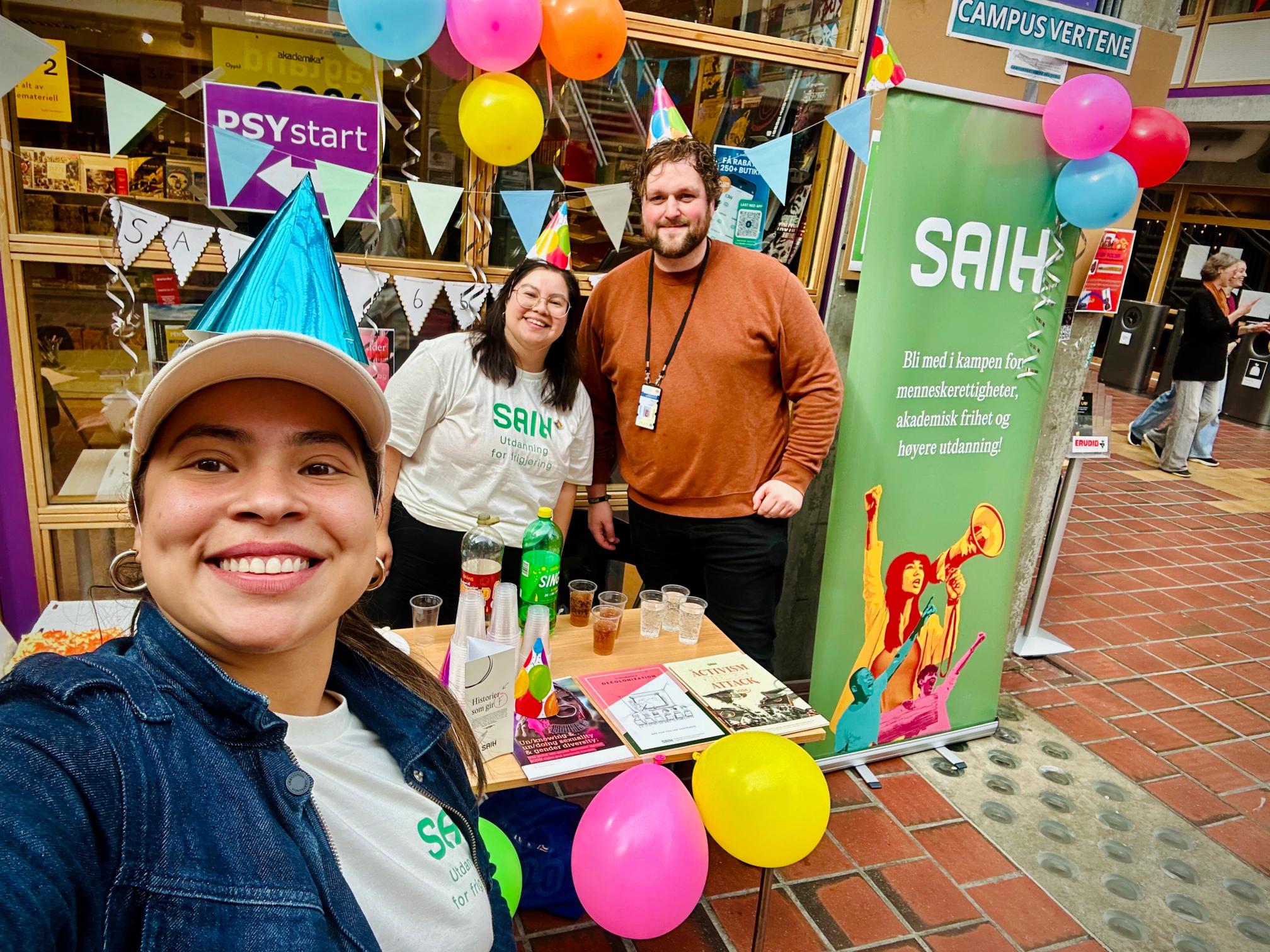 A selfie of three smiling people at a SAIH event booth decorated with balloons and a green banner.