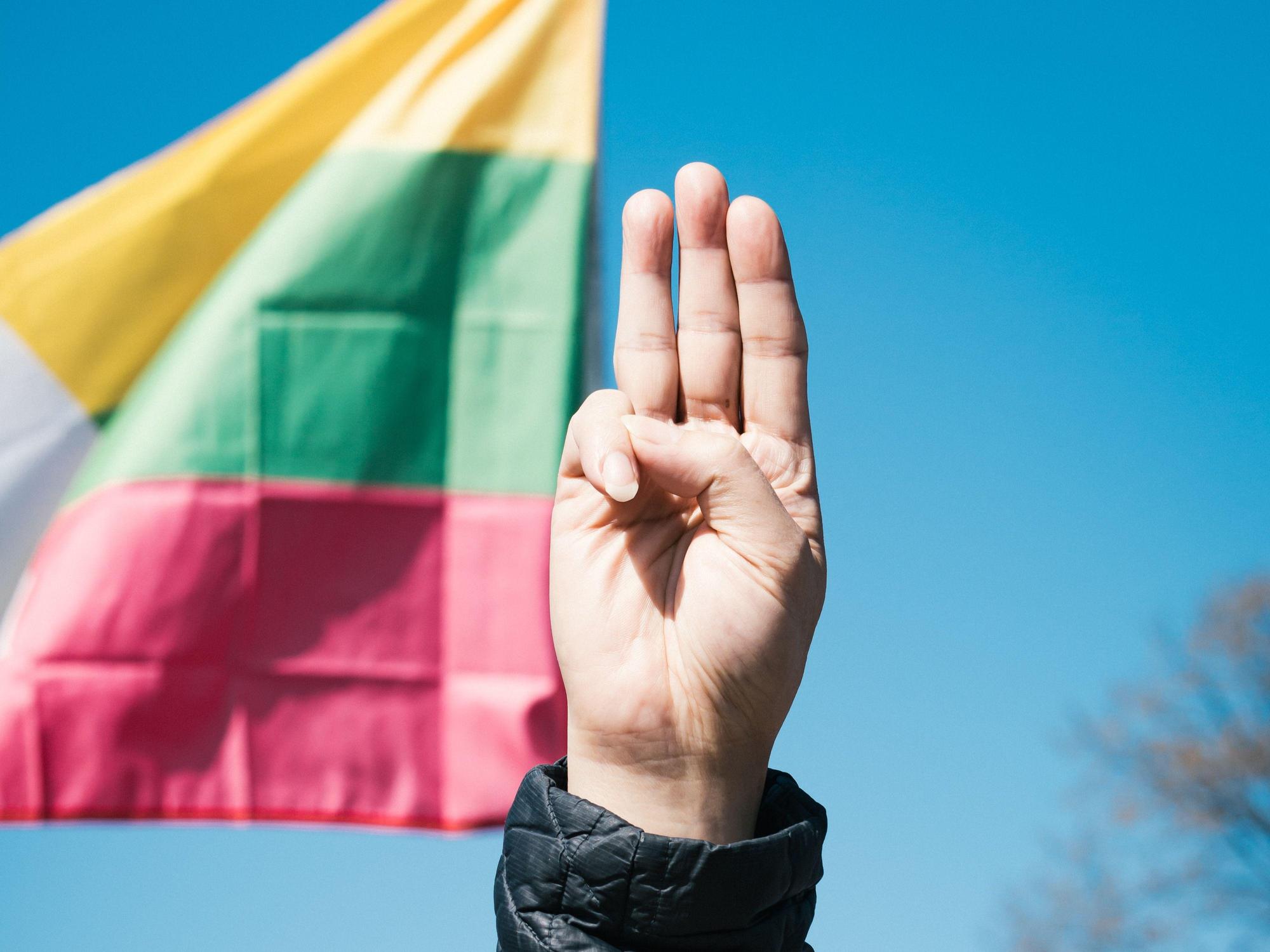 A hand showing three fingers, with a Burmese flag in the background.