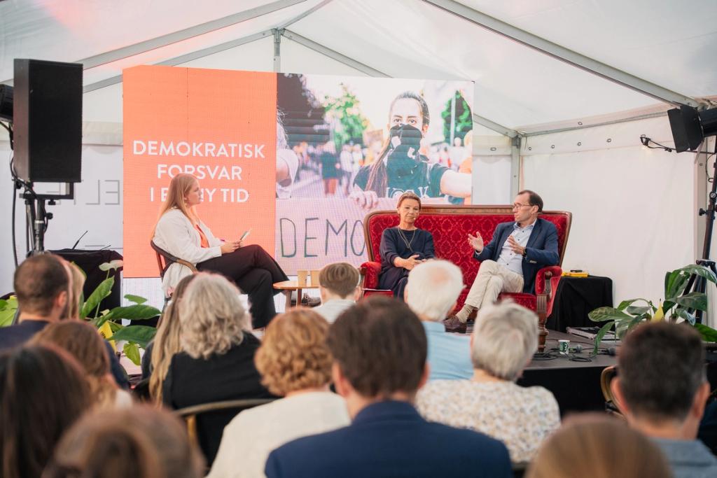 A panel discussion with three people on a stage in a white tent, two seated on a red sofa, addressing an audience. A large screen behind them displays 'DEMOKRATISK FORSVAR I EN NY TID' and an image.