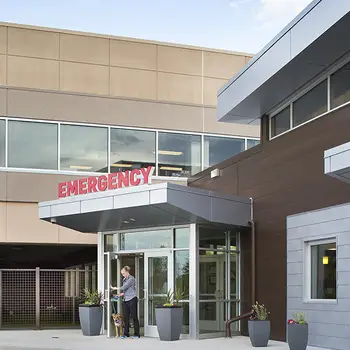 Woman with a dog exits the door of an emergency department with glass doors and a red "EMERGENCY" sign above.