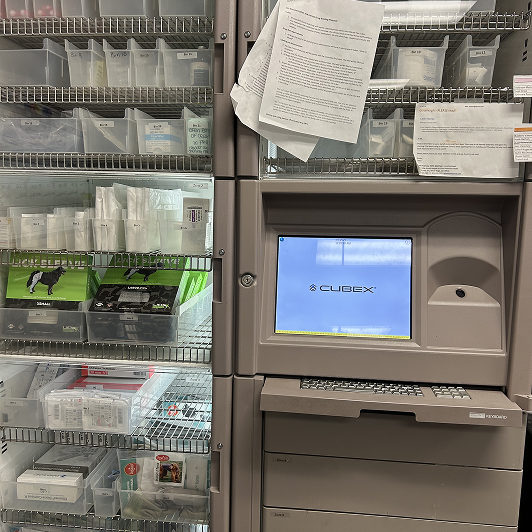 A veterinary hospital supply area with shelves of medical supplies and a CUBEX electronic cabinet featuring a screen and keyboard.