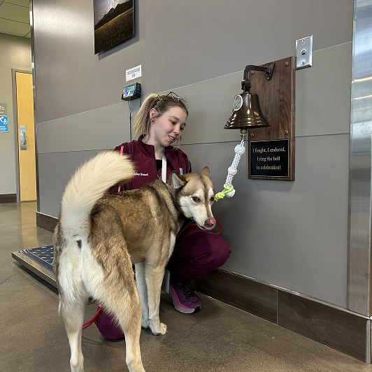 A woman in maroon scrubs with a Husky dog by a mounted bell.