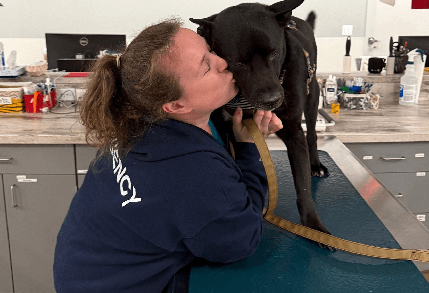 A vet with a blue jacket reading "Emergency" across the back kisses a large black dog in a clinical setting.