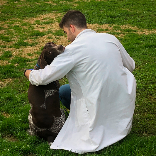 A man in a white lab coat kneels with his arm around a brown dog looking back, as seen from behind.