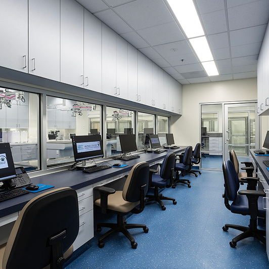 A modern laboratory with workstations and computers along a wall, blue speckled flooring, and white overhead cabinets.