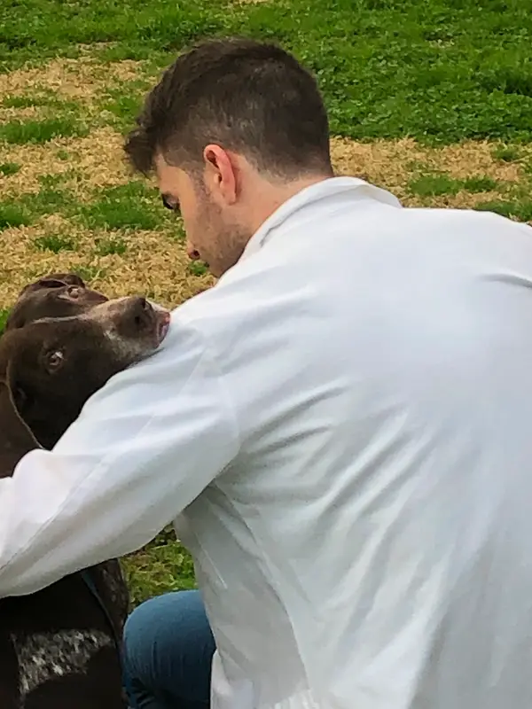 A man in a white lab coat embraces a brown dog on a grassy lawn, as seen from the back.