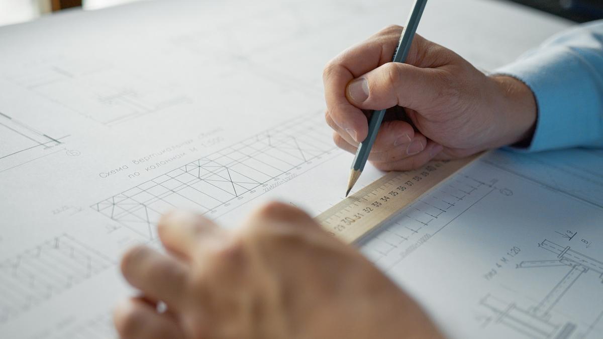 Nærbilde av arkitekttegninger og to hender, venstre hånd holder en linjal over tegningen og høyre hånd holder en blyant.