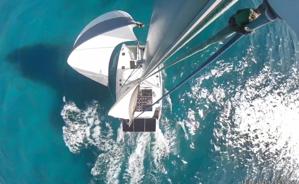 Aerial shot of a catamaran sailing through clear turquoise water with sails full.