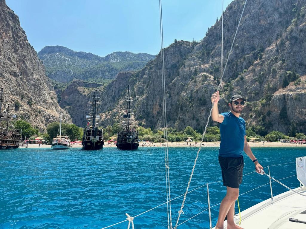 Sailor standing on bow of sailboat in clear blue water with steep cliffs behind.