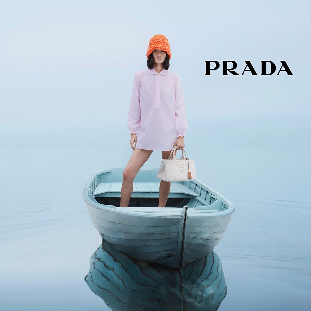 Prada ad showing a model with a white handbag, pink outfit and orange hat standing in a pale blue boat.