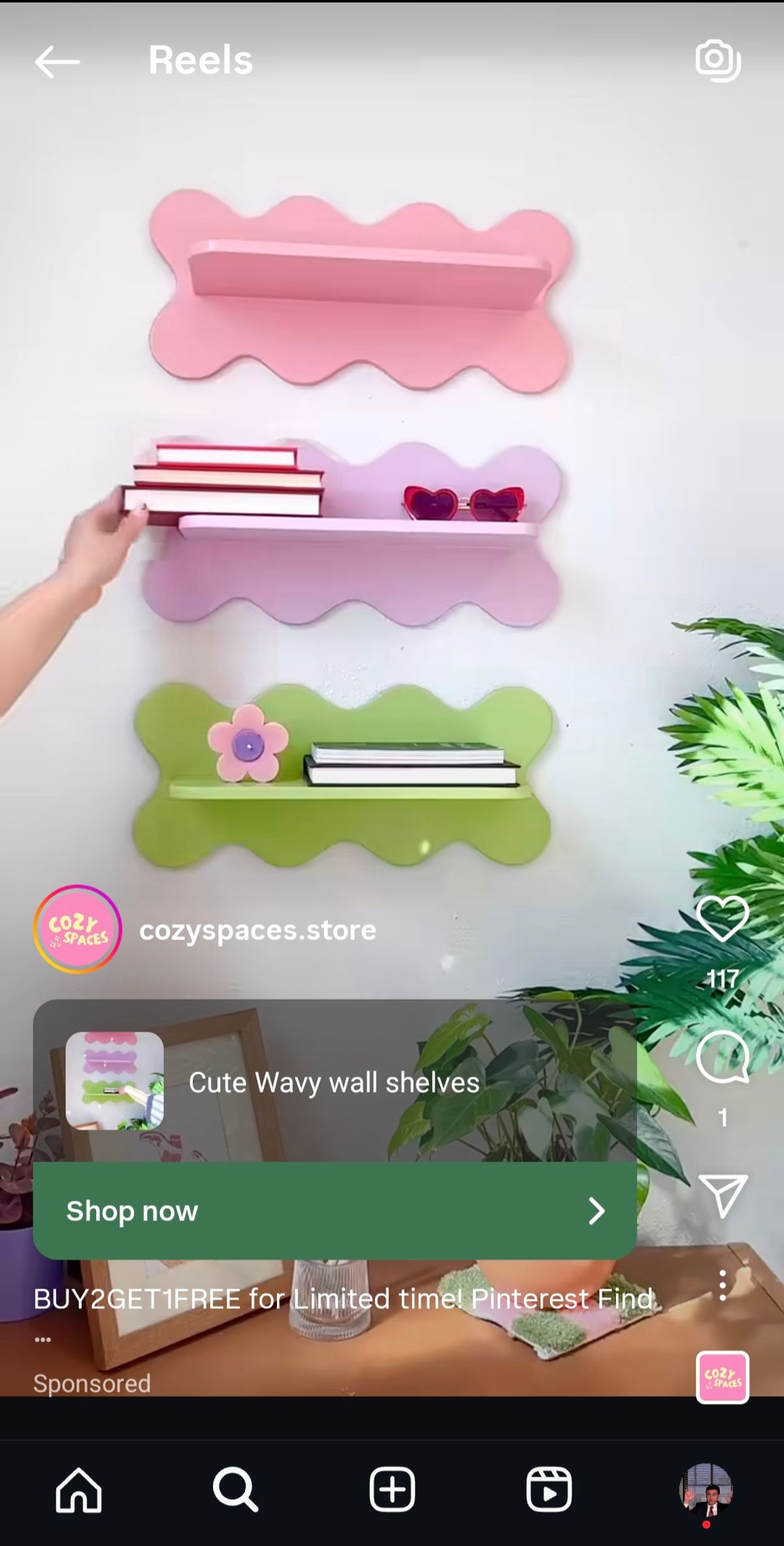 A still frame from an Instagram Reel Ad by cozyspaces.store showing a hand placing items on three wavy, colorful wall shelves: one pink, one lavender, and one lime green. The ad promotes 'Cute Wavy wall shelves' and a 'BUY 2 GET 1 FREE' offer. The Reel format allows the ad to blend with organic content and features a 'Shop now' call-to-action button for direct conversion.