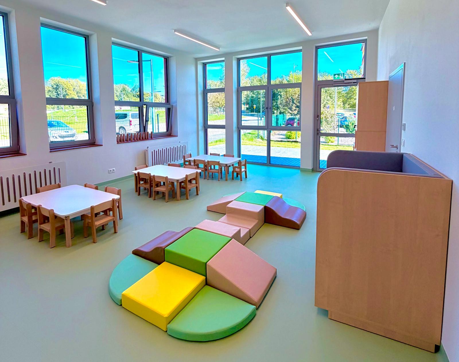 Przestronna sala z dużymi oknami, stolikami dla dzieci i miękkimi klockami