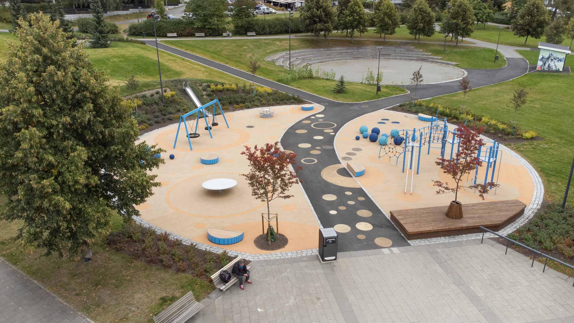 Aerial view of Romsaas Park playground featuring swings, slides, climbing structures, and colorful play areas. The park is surrounded by green lawns, pathways, and trees, providing a vibrant recreational space for families and children.