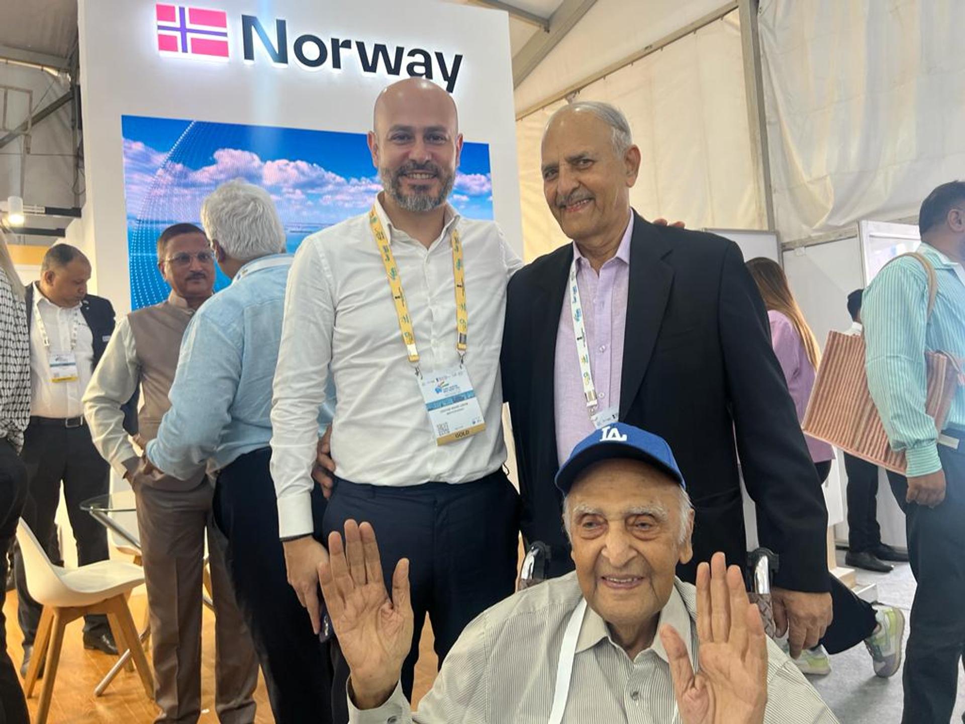 Three men smiling in front of a Norwegian pavilion