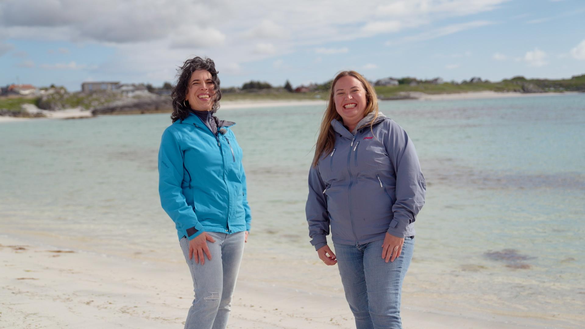 Two women in blue jackets on a white sand beach with turquoise water behind them