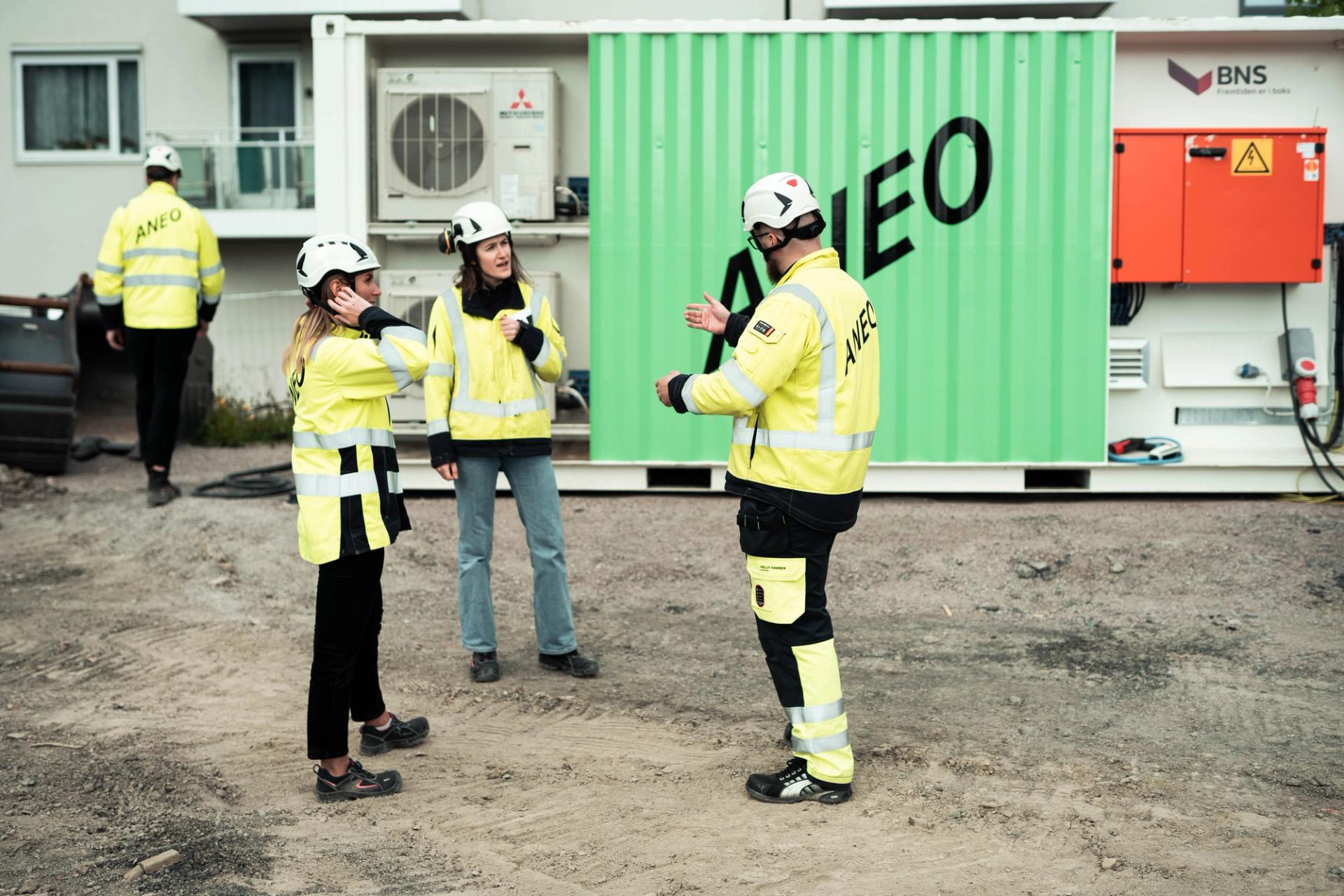Four people in yellow work jackets and hard hats in front of a green charger container