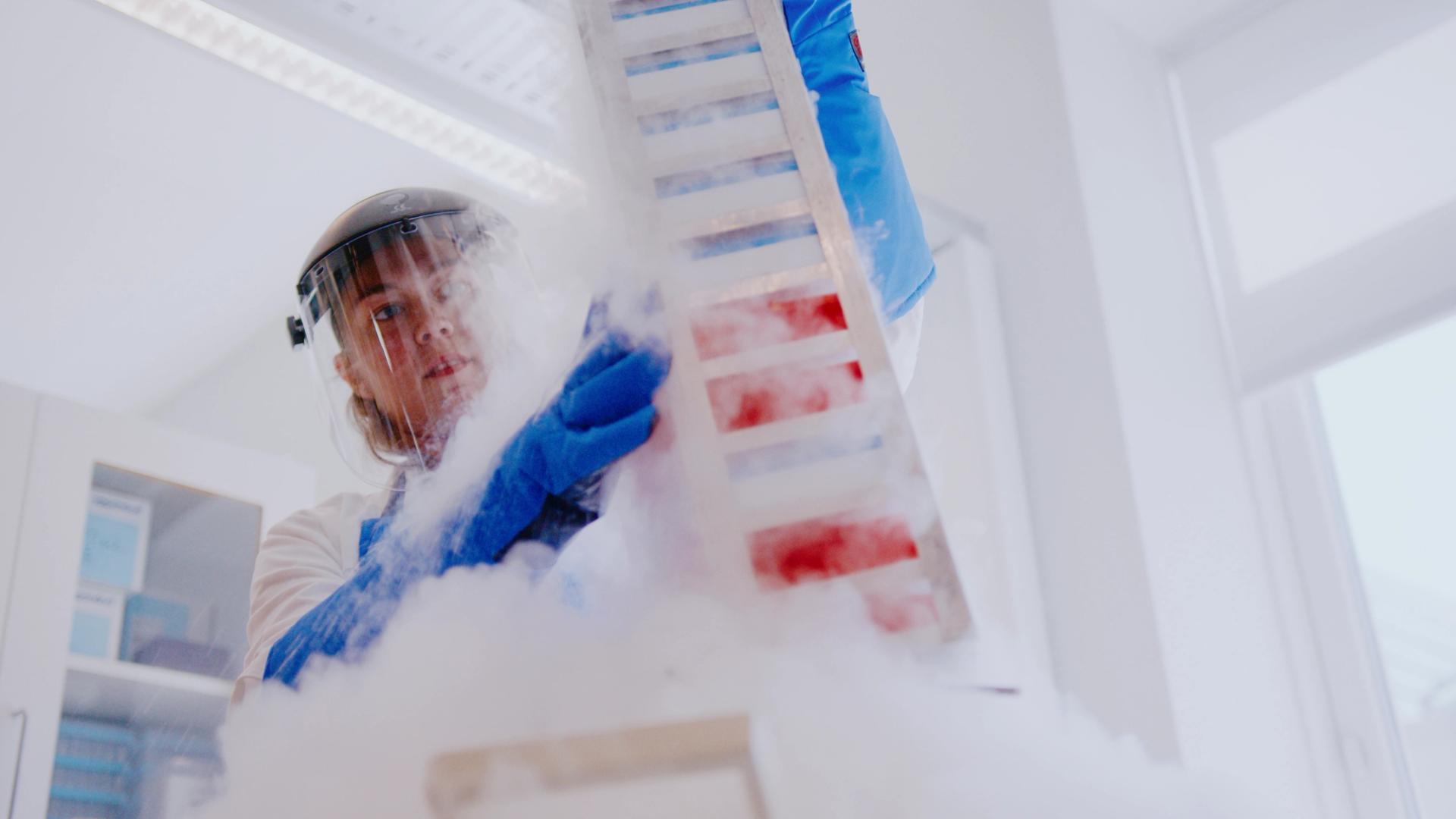 Woman in a lab coat and protective gloves and mask lifting a set of small drawers out of cold smoke.