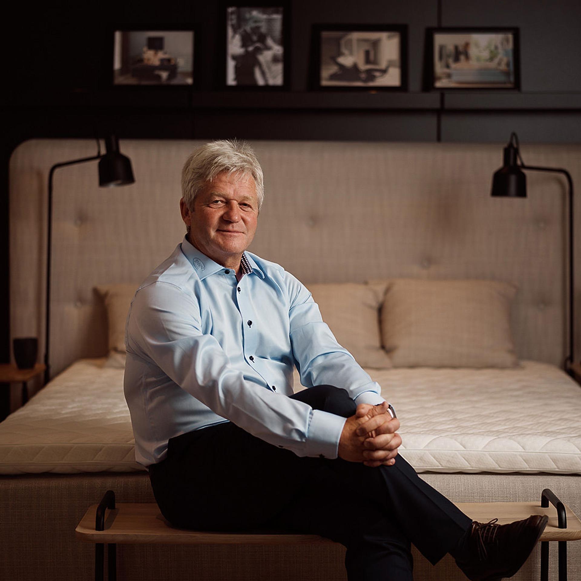 A man, Jan Trygve Jensen, smiling to the camera while sitting on a bench infant of a bed.