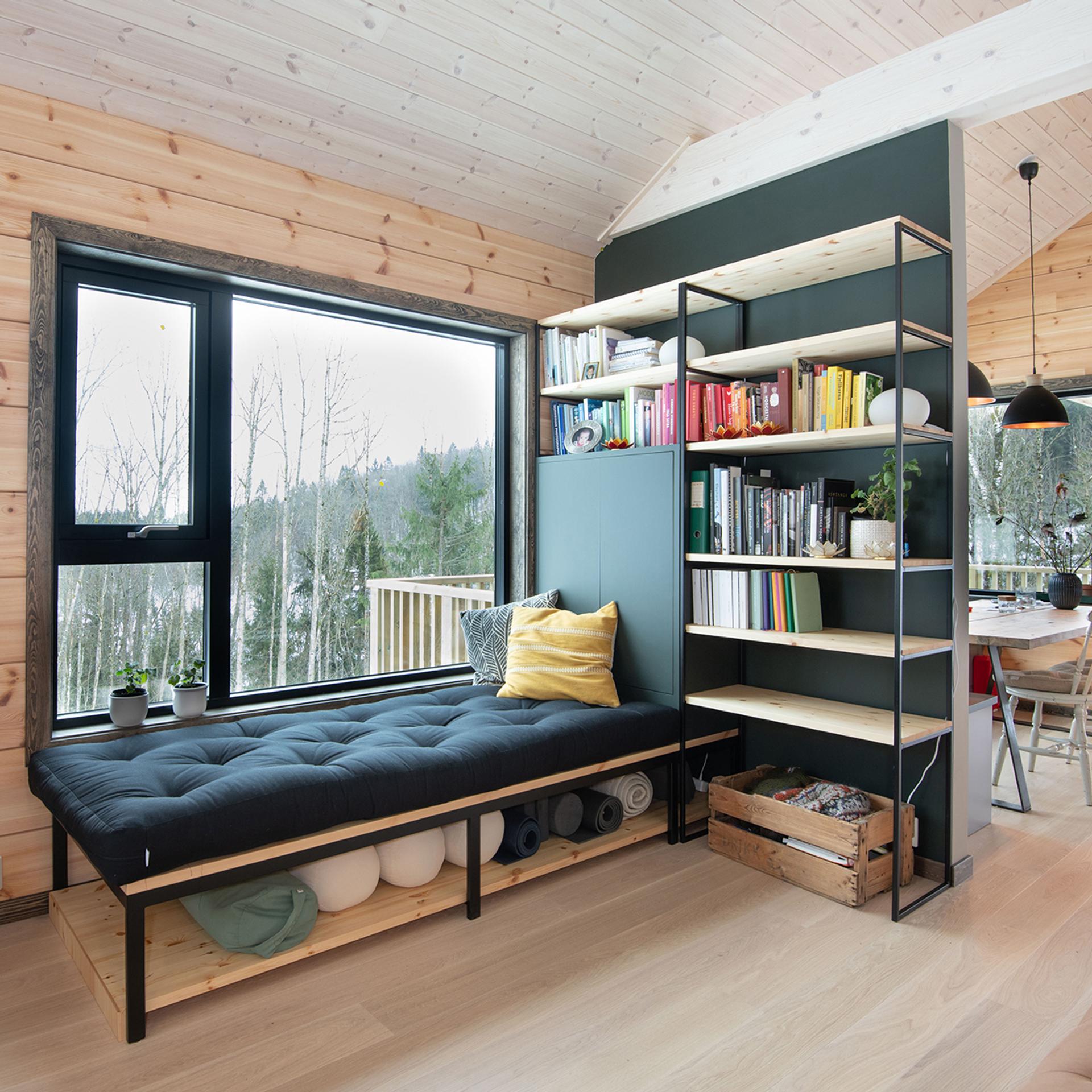 Reading nook with a navy daybed, forest-view window and a bookshelf, in a timber cabin.