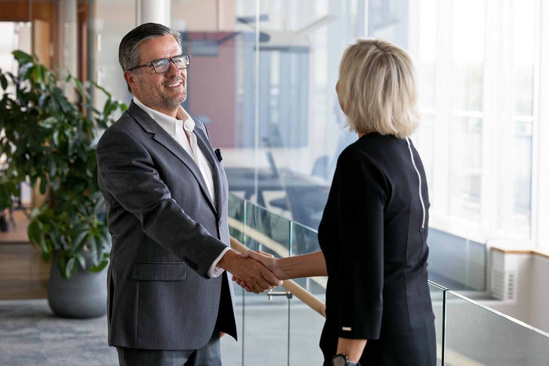 Man with gray hair in gray blazer and glasses greeting a blond woman in a black dress
