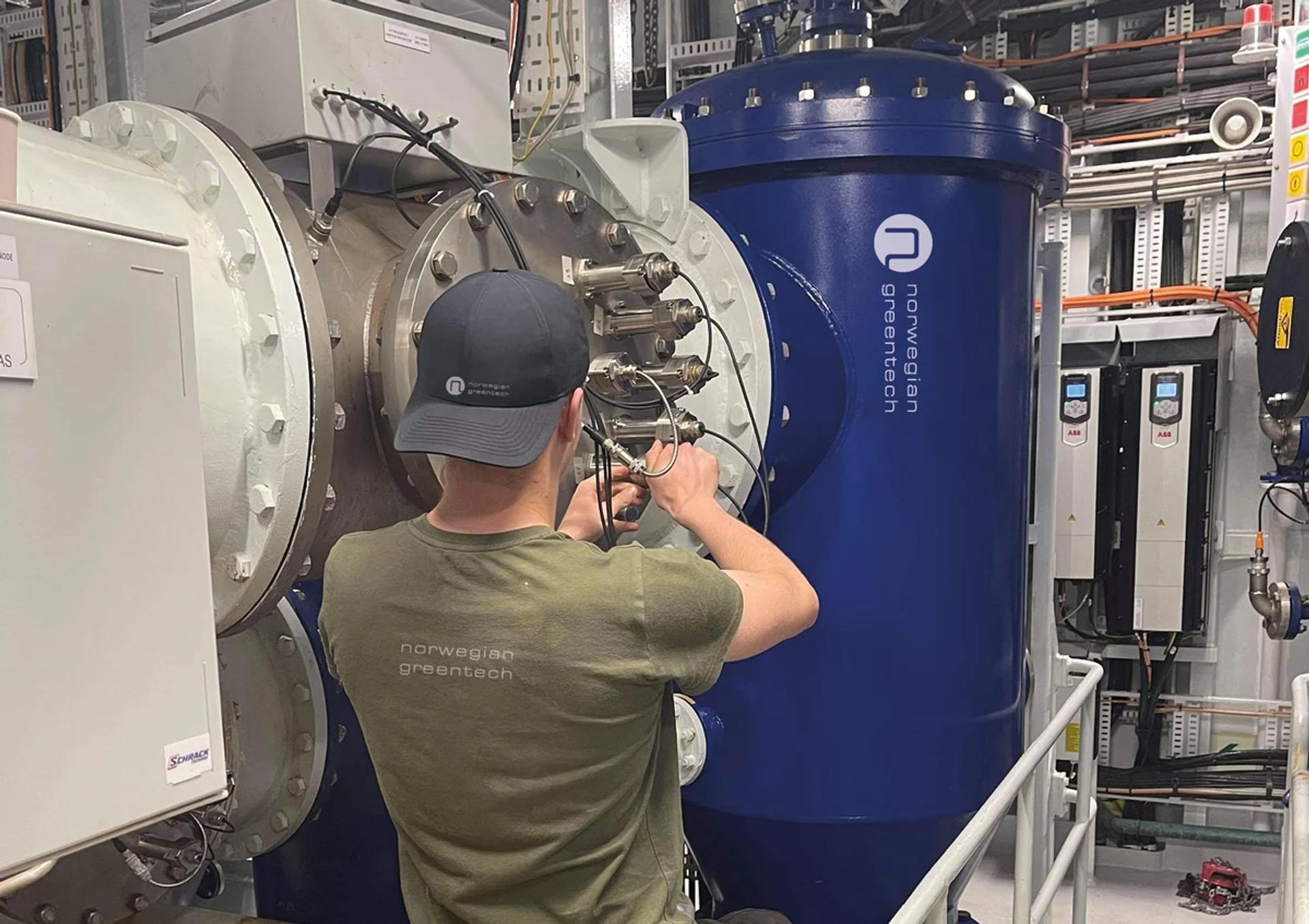 man in a green shirt and black baseball cap attaching wires to a ship component
