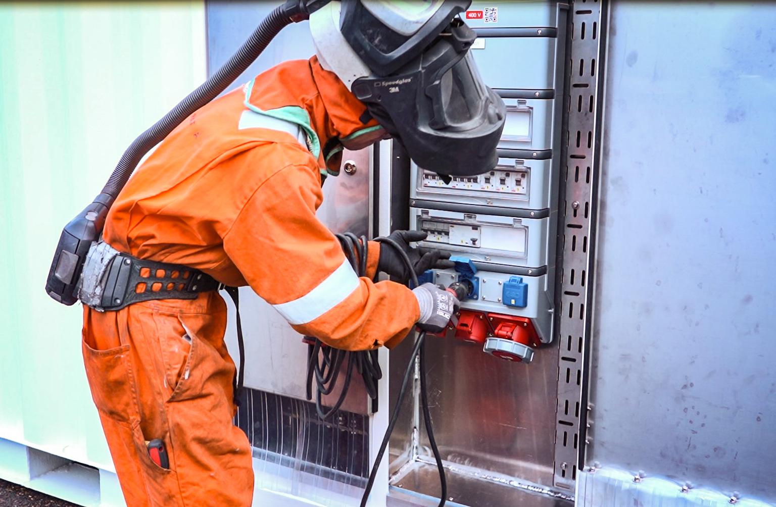 Man in an orange worksuit and helmet plugging a cable into a mobile battery