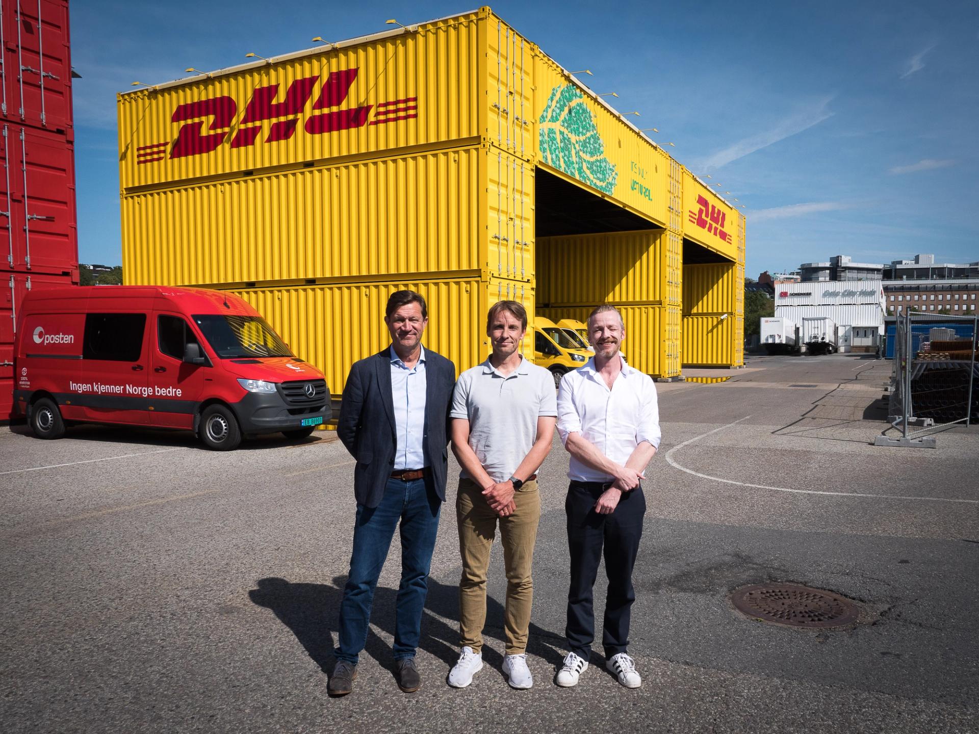 Three white men in white shirts in front of a huge yellow DHL garage