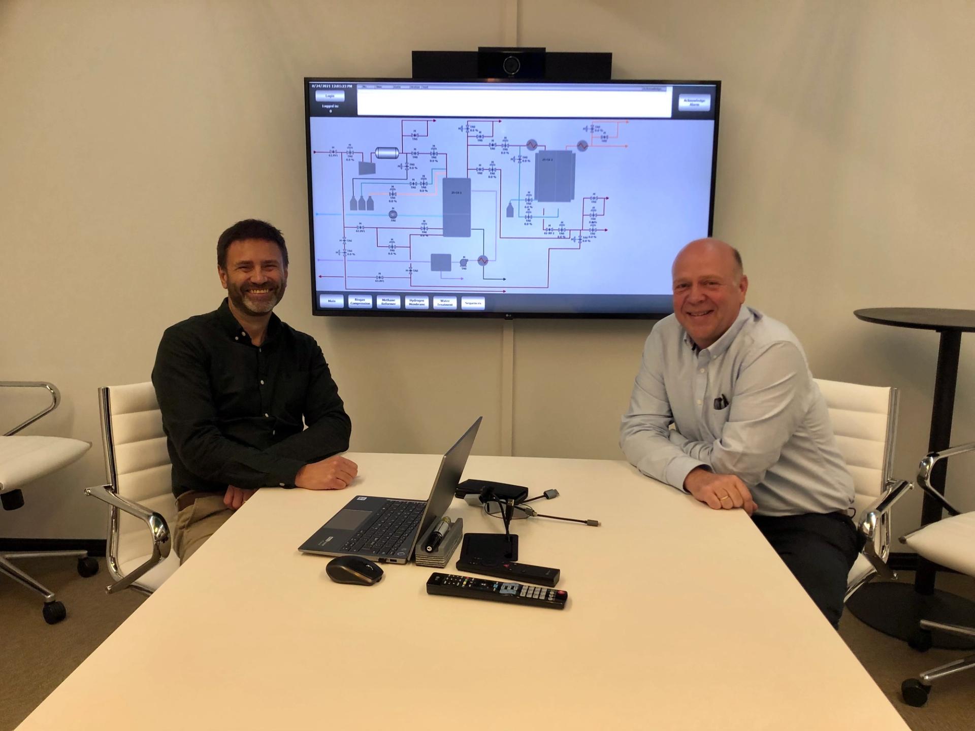 two smiling men at a desk in front of a screen with a schematic