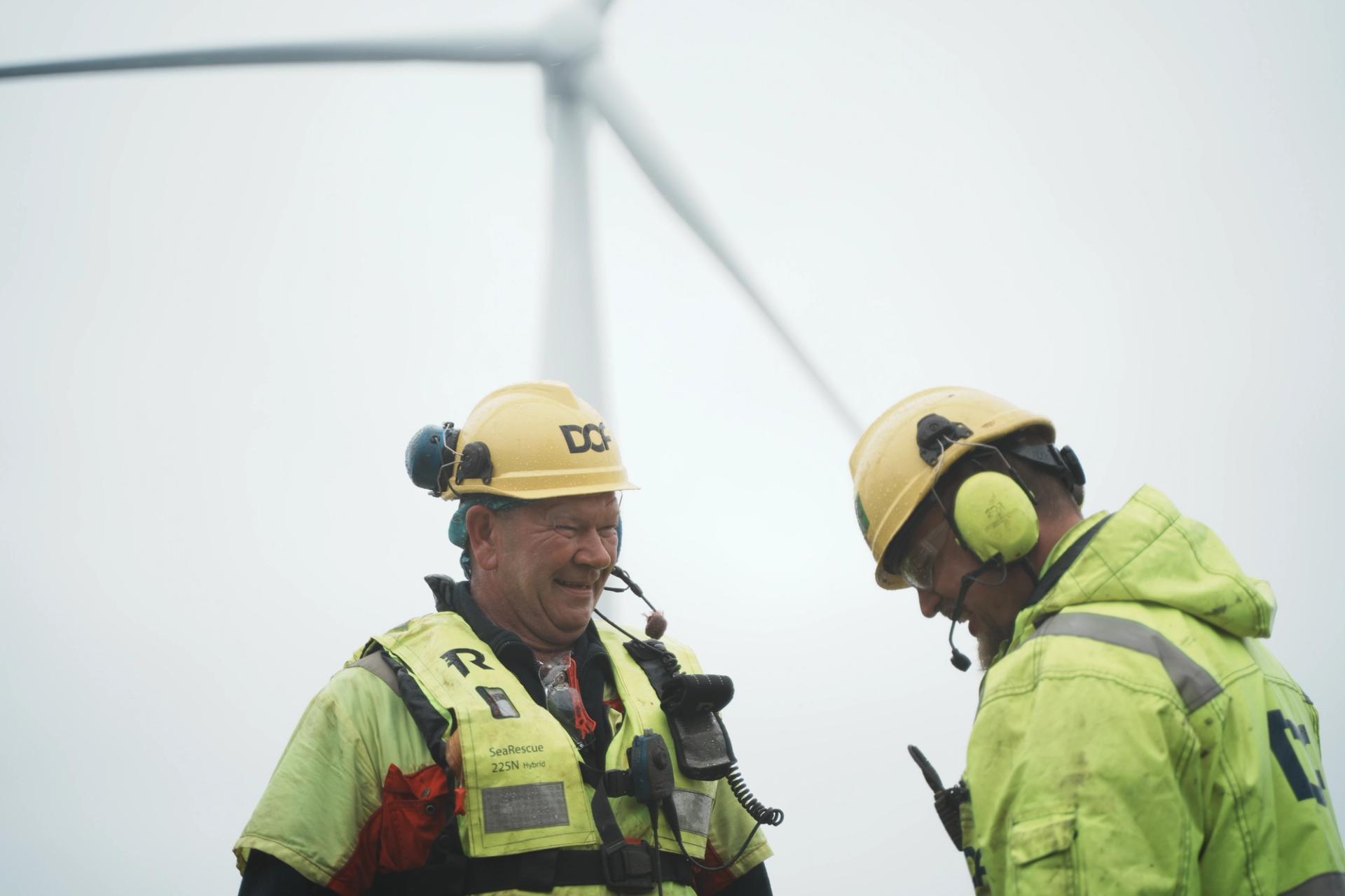 Two men in yellow safety gear and hard hats in front of an offshore wind turbine