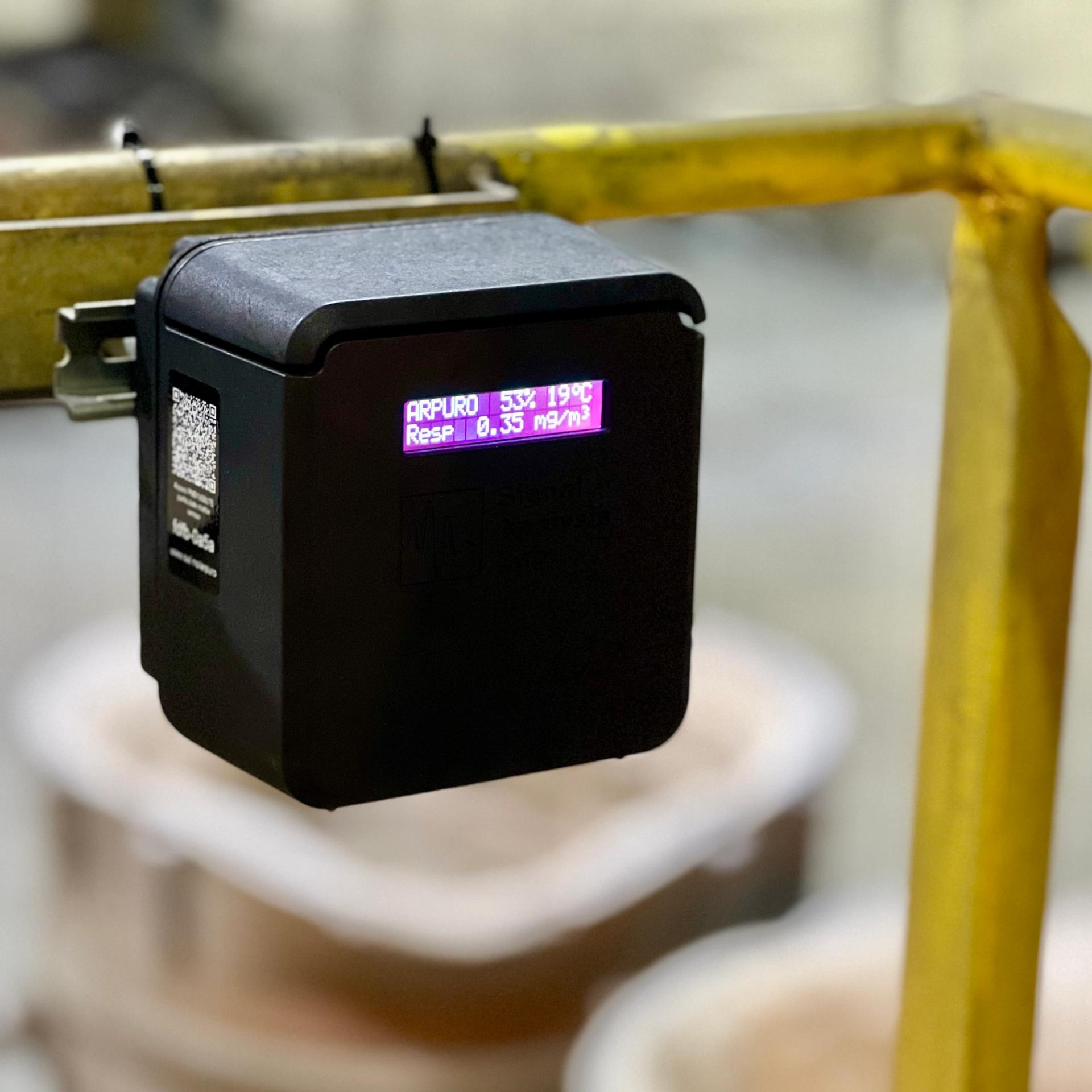Indoor industrial dust monitor housed in a black box hanging from a yellow pipe
