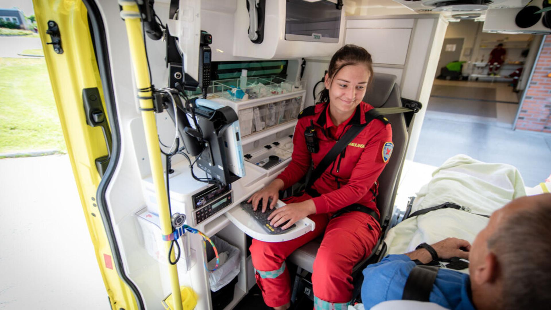 Female paramedic in a red suit working at a tablet and male patient on a gurney