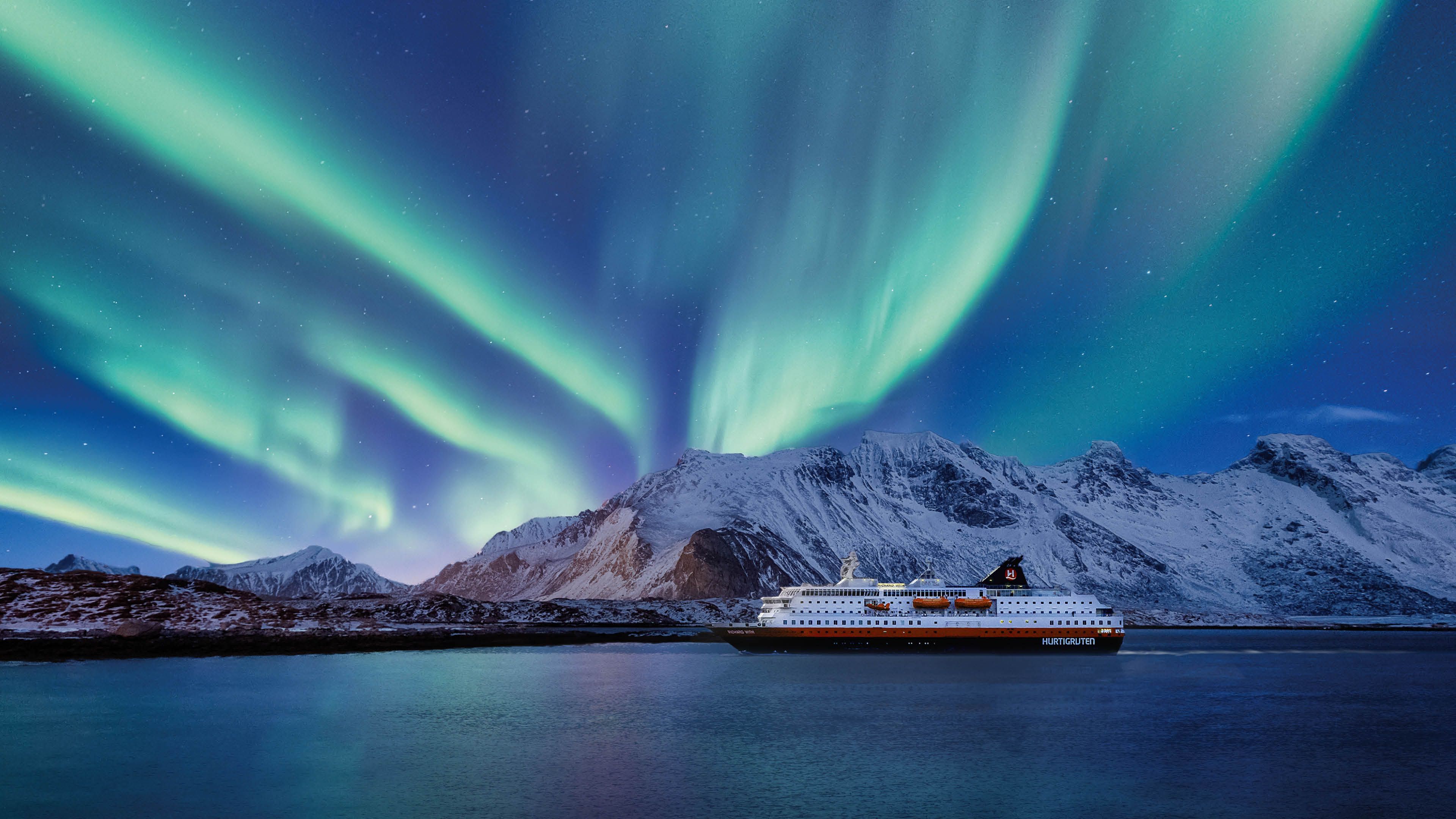 A Hurtigruten ship sails under the Northern Lights in Norway, with snow-capped mountains in the background