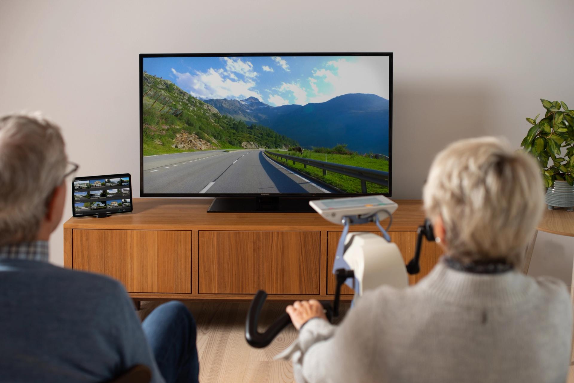 Two grey haired people on a stationary bike watching a road on a TV