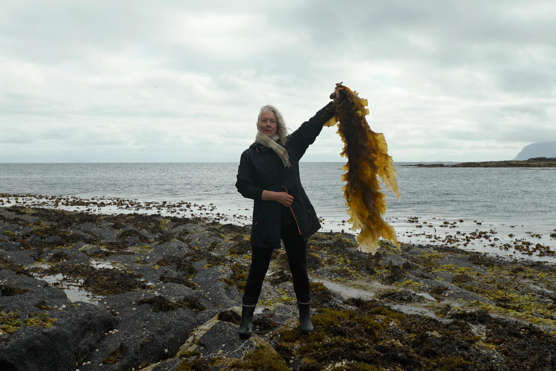Blond woman standing on the shore holding up brown seaweed