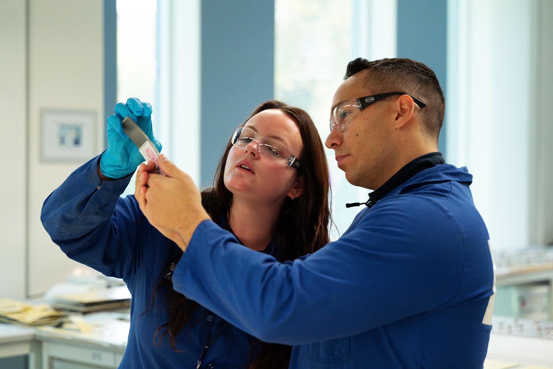 Two people in blue lab coats and protective goggles looking at a test tube
