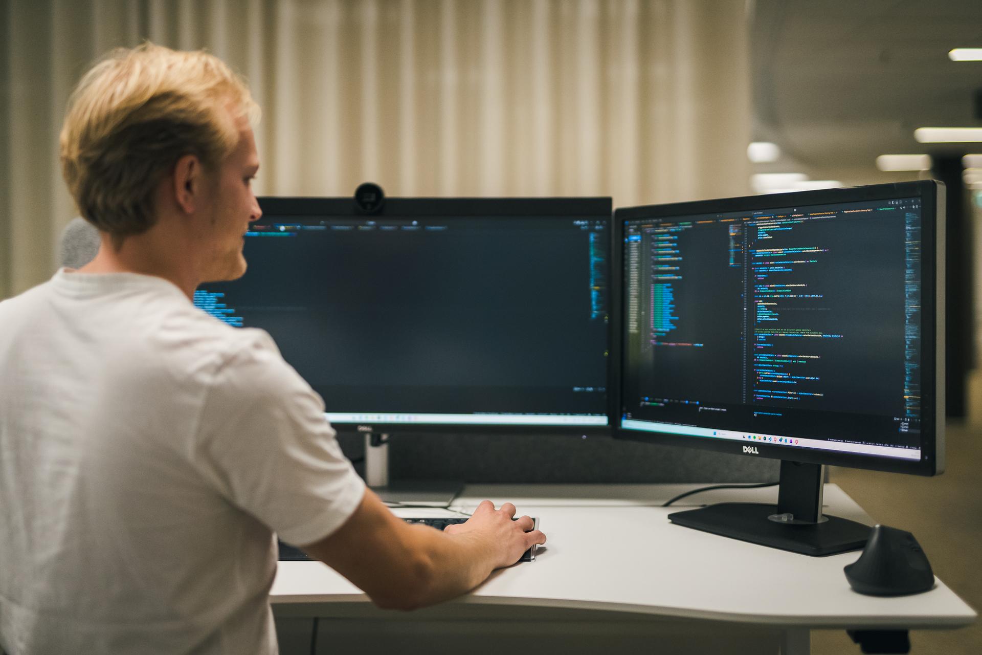 Man in a white t-shirt at a desk looking at two computer screens