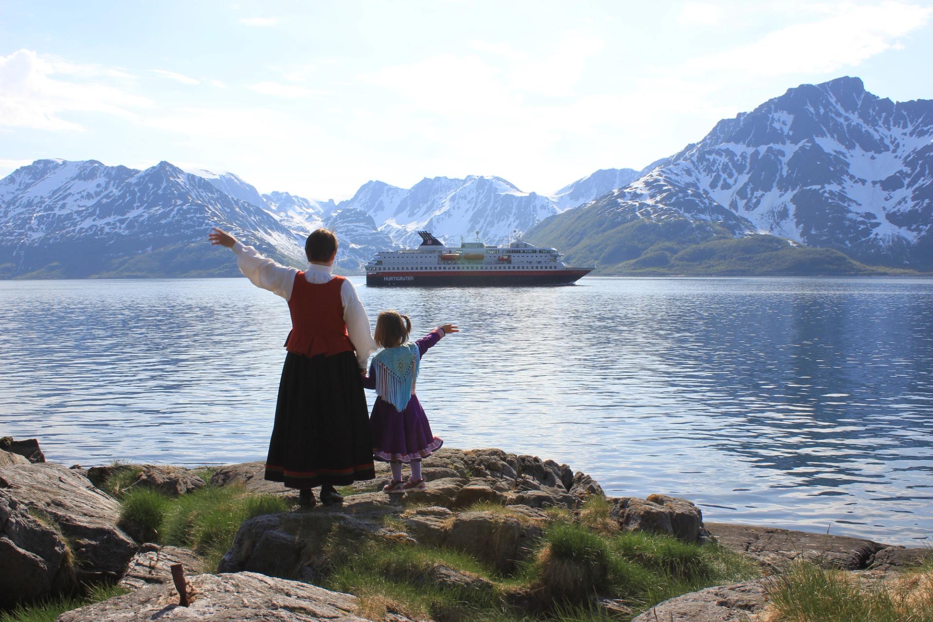 Two people in national costumes stand on a rocky shore, waving to a Hurtigruten ship sailing by in a fjord surrounded by snow-capped mountains