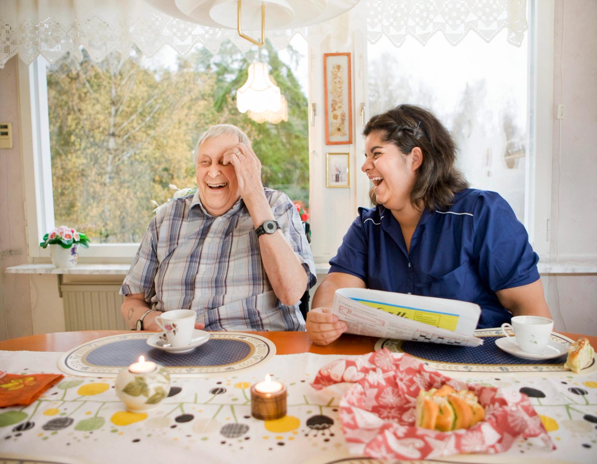 Old man and care giver drinking coffee at a table and laughing