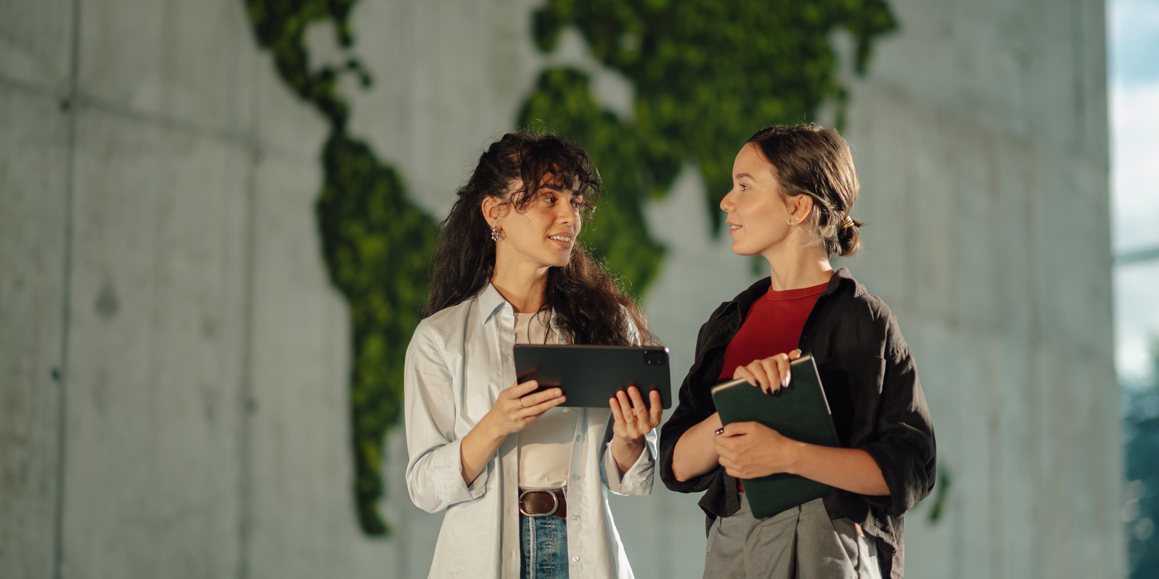 To forretningskvinner i diskusjon foran en steinvegg med et grønt verdenskart av blader på seg.