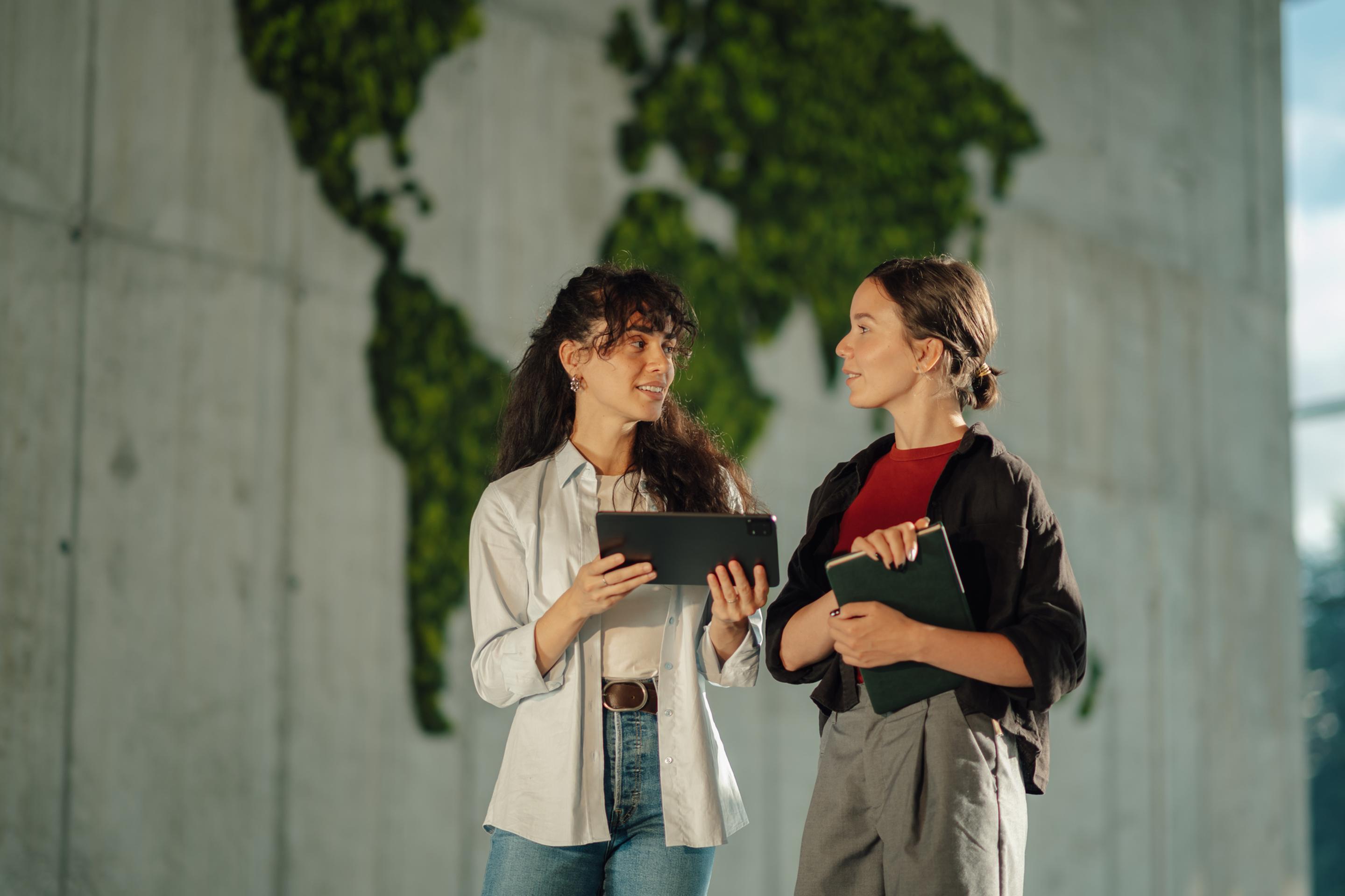 To forretningskvinner i diskusjon foran en steinvegg med et grønt verdenskart av blader på seg.