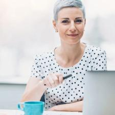 A woman is sitting in front of her computer with a cup of coffee, smiling.