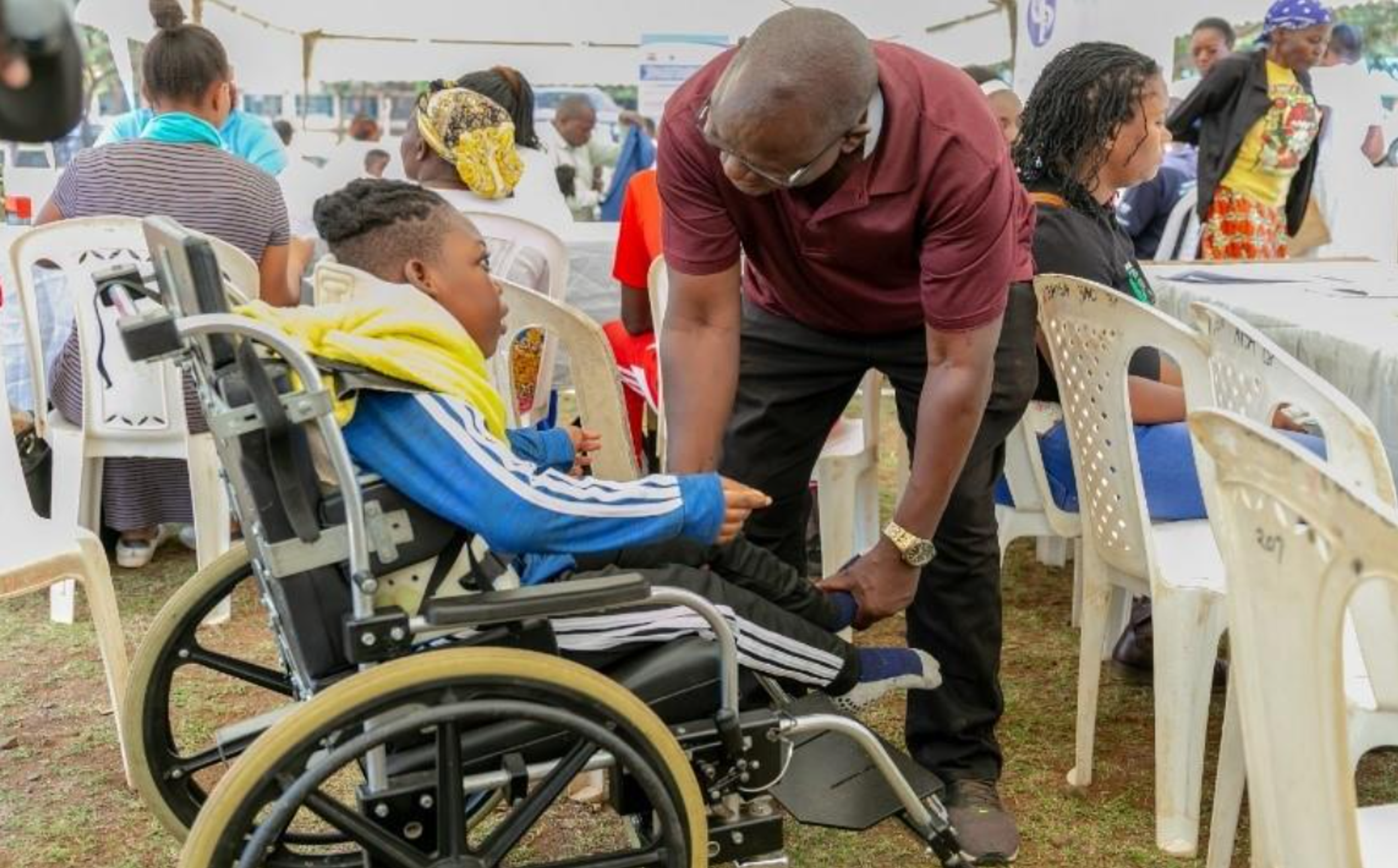 A child seated in a wheelchair receives assistance from an adult who is leaning down to adjust the child's foot or leg support. They are outdoors under a tent, surrounded by several people seated on plastic chairs at tables.