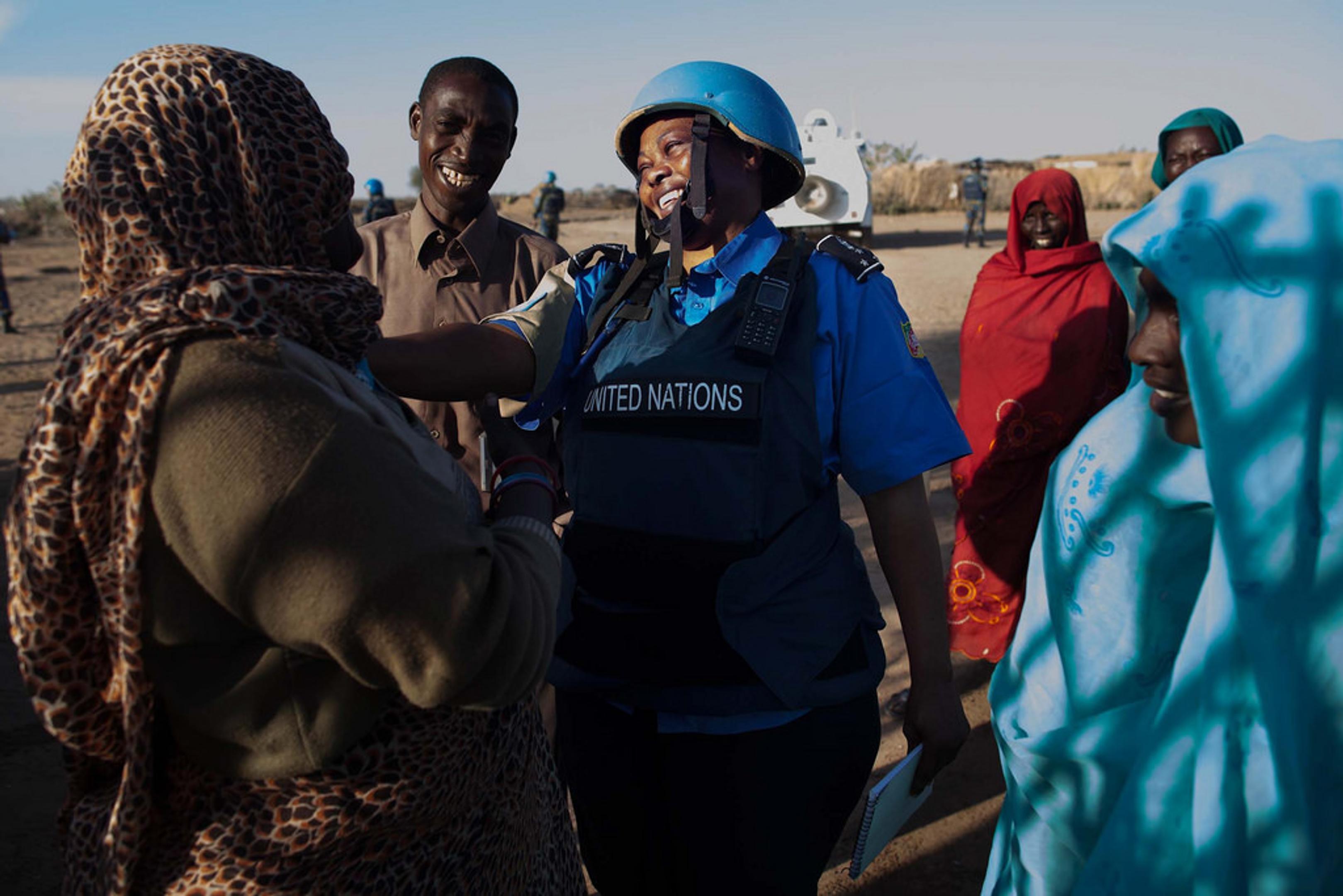 A woman wearing a blue helmet and protective UN vest with speaks with a group of people in traditional clothing in an outdoor, desert-like area. Several individuals stand nearby, appearing to engage in the conversation or observe the interaction.