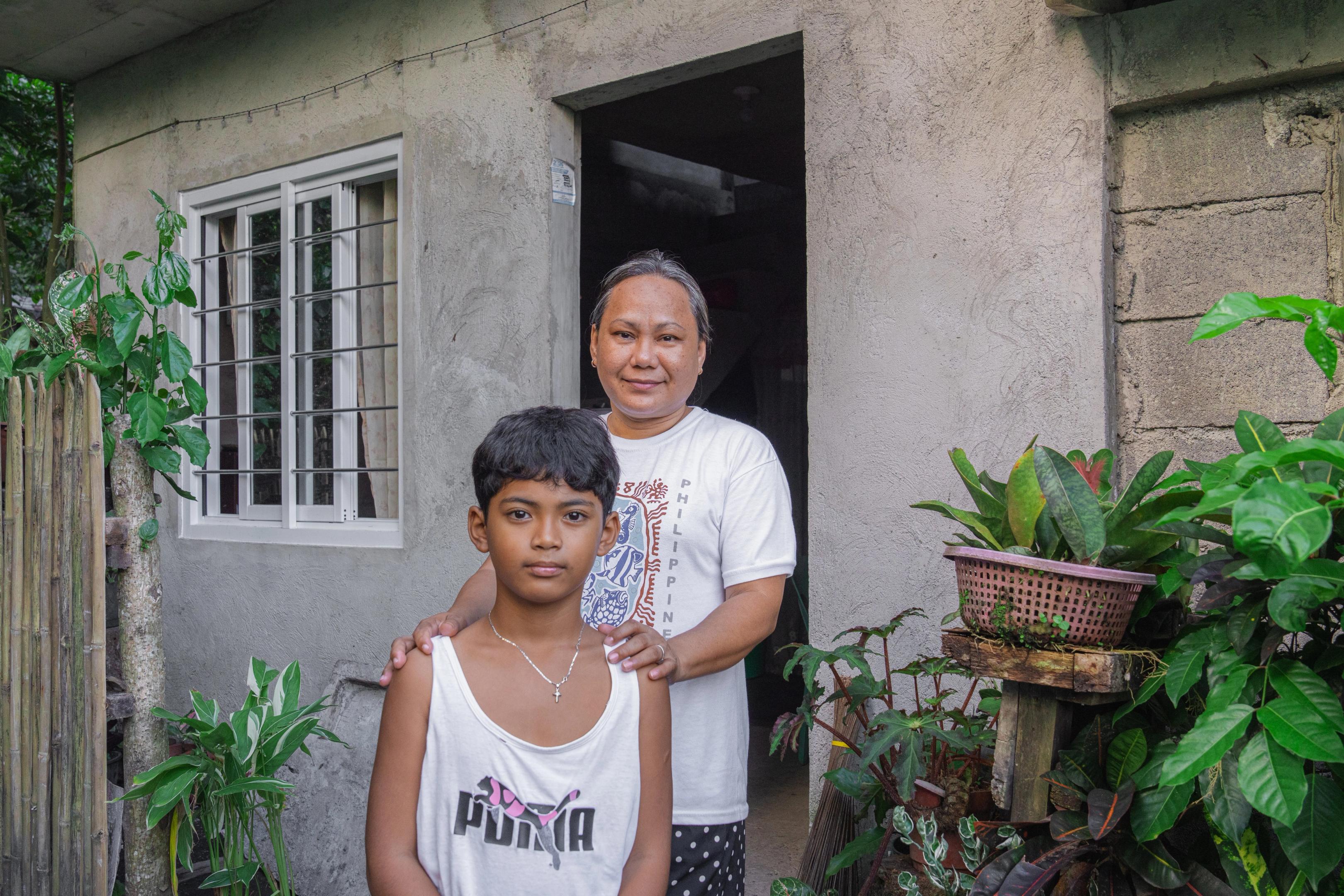 An adult stands behind a child outside a small concrete house, with the adult’s hands resting on the child’s shoulders. The house has a window with white bars, an open doorway, and a variety of green plants arranged in pots near the entrance.